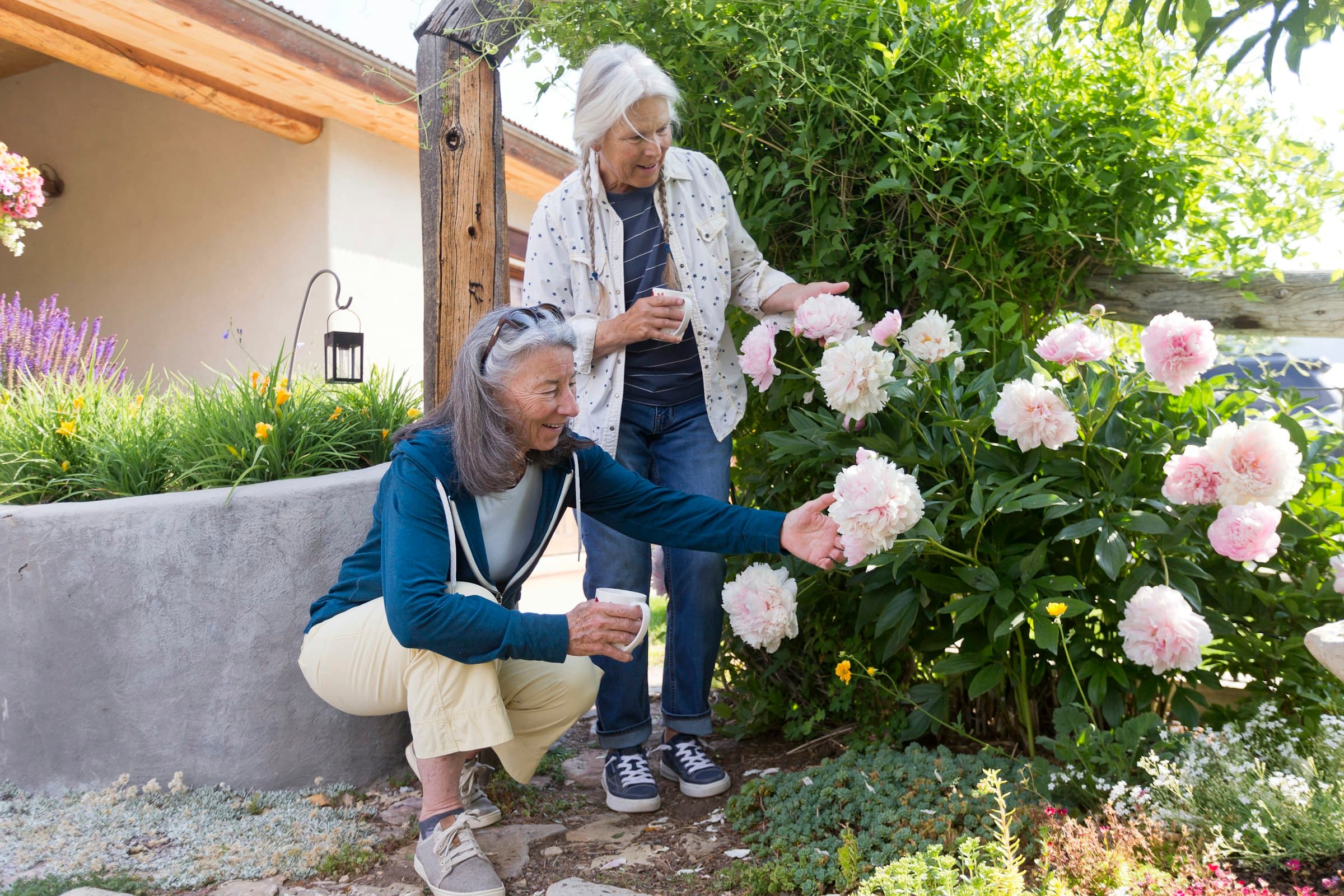 Two women in a garden, one crouching to touch a pink peony, the other standing behind her among the flowers.