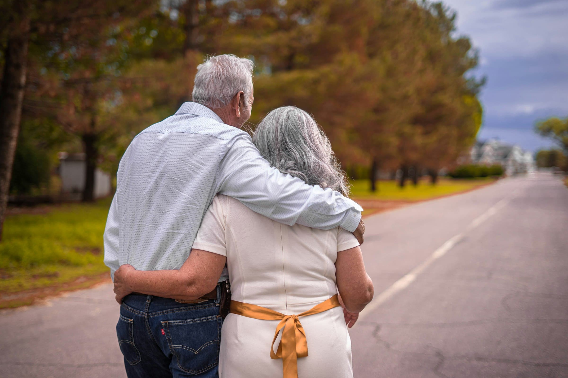 An older man and woman with grey hair, walking away down a tree-lined road with his arm around her shoulders.
